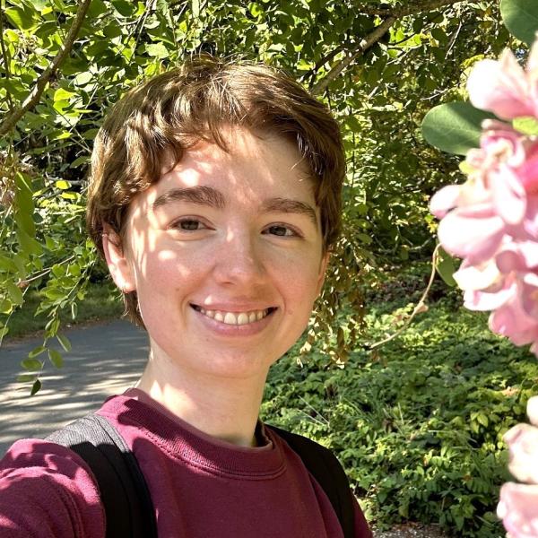 Person with blonde hair and a red sweatshirt smiles at the camera, with a flower in the foreground.
