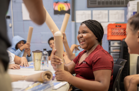A smiling girl with black hair holds a brown tube surrounded by classmates