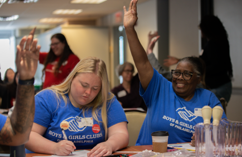 A woman with dark hair eagerly raising her hand next to a woman with blonde hair who is writing