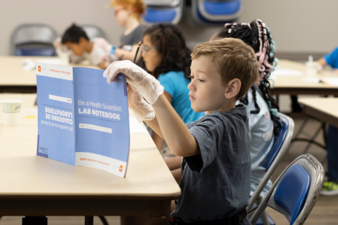 A blonde boy looks at a blue lab notebook while sitting at a table
