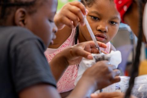 A young girl and two other learners lean close together, with one learner holding a funnel containing filter paper and soil, and the other investigating with a pipette.
